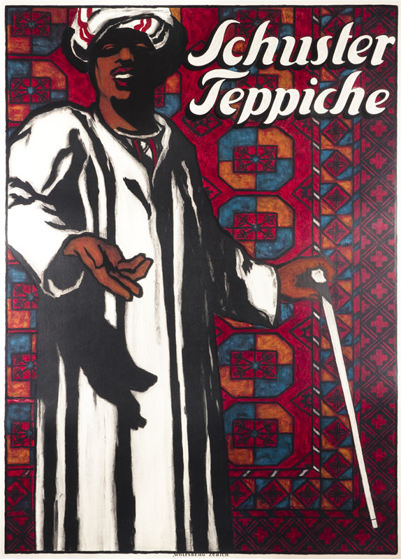 Middle Eastern man stands in front of rugs; white, burgundy, orange, black